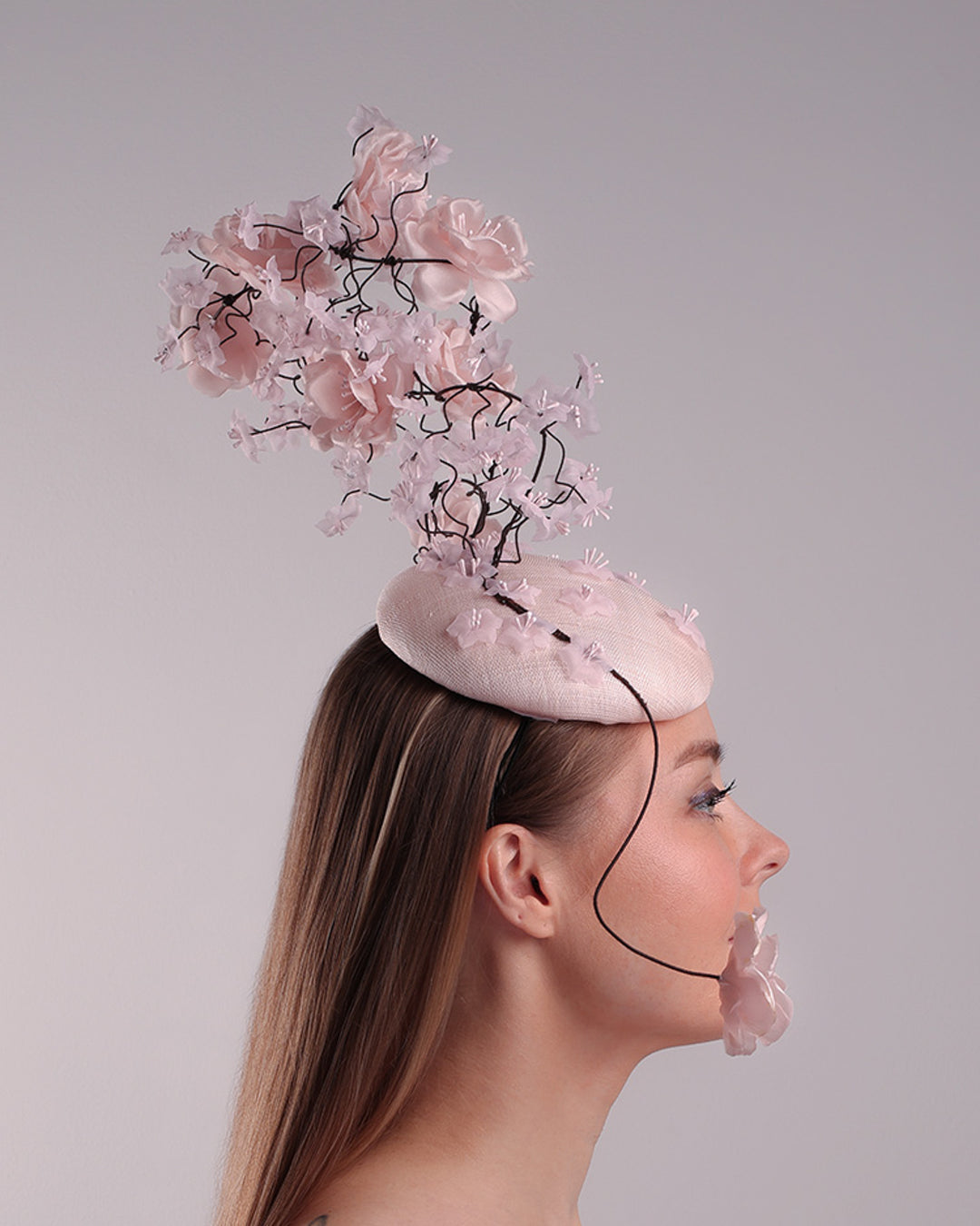 Model is showcasing the right side of the button shaped pink fascinator with sinamay base decorated with tree like wire branches embellisehd with custom made pink silk flowers. The pink button fascinator is placed on the right front side of the face and the wire braces go up towards the left side of the button base while a single branch with one rose falls on the right side of the face.