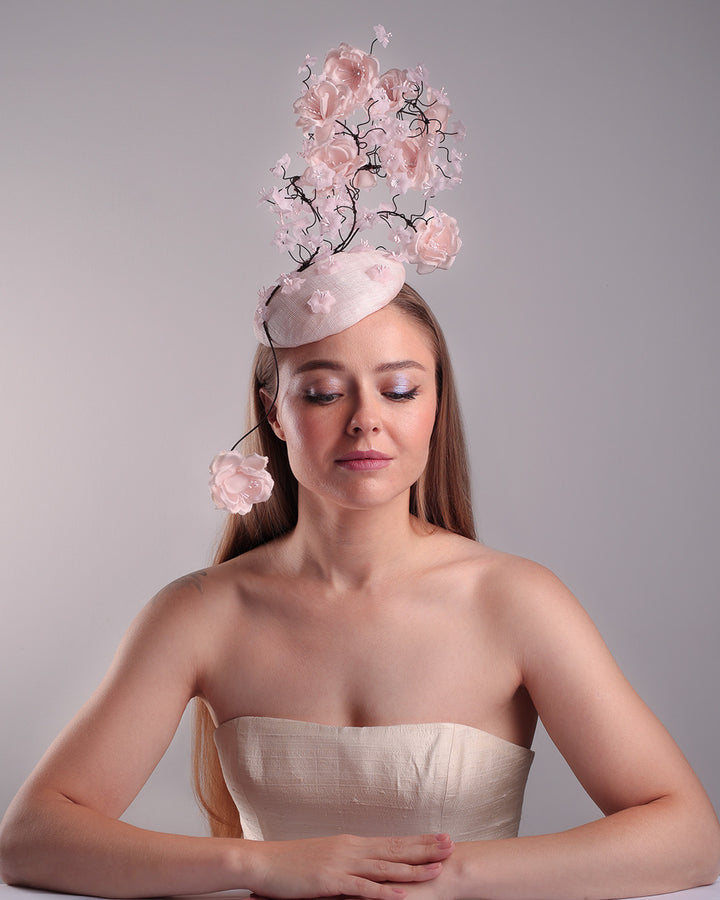 Model is showcasing the ront of the button shaped pink fascinator with sinamay base decorated with tree like wire branches embellisehd with custom made pink silk flowers. The pink button fascinator is placed on the right front side of the face and the wire braces go up towards the left side of the button base while a single branch with one rose falls on the right side of the face.