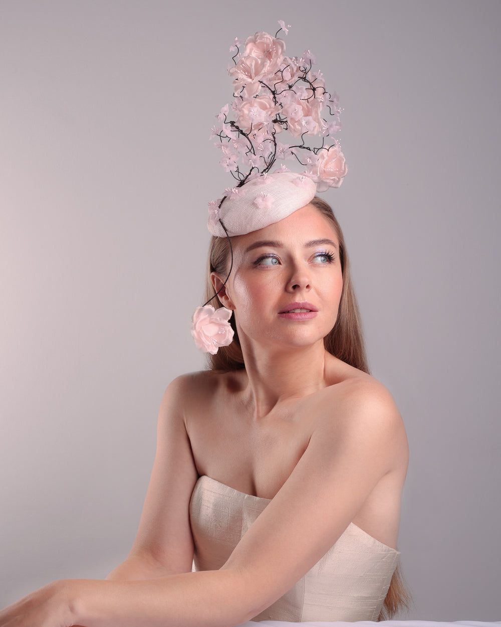 Model is showcasin the right front angle of the button shaped pink fascinator with sinamay base decorated with tree like wire branches embellisehd with custom made pink silk flowers. The pink button fascinator is placed on the right front side of the face and the wire braces go up towards the left side of the button base while a single branch with one rose falls on the right side of the face.