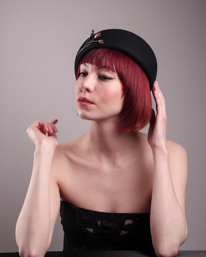 The female model is wearing a wool beret-style hat, showcasing the left front view it. The black hat is decorated with a real gold-plated brass metal branch motif adorned with crystal-tipped buds. The side beret is higher on the left side and shorter on the right side, and is suggested to be worn tilted. Designed by Merve Bayindir.
