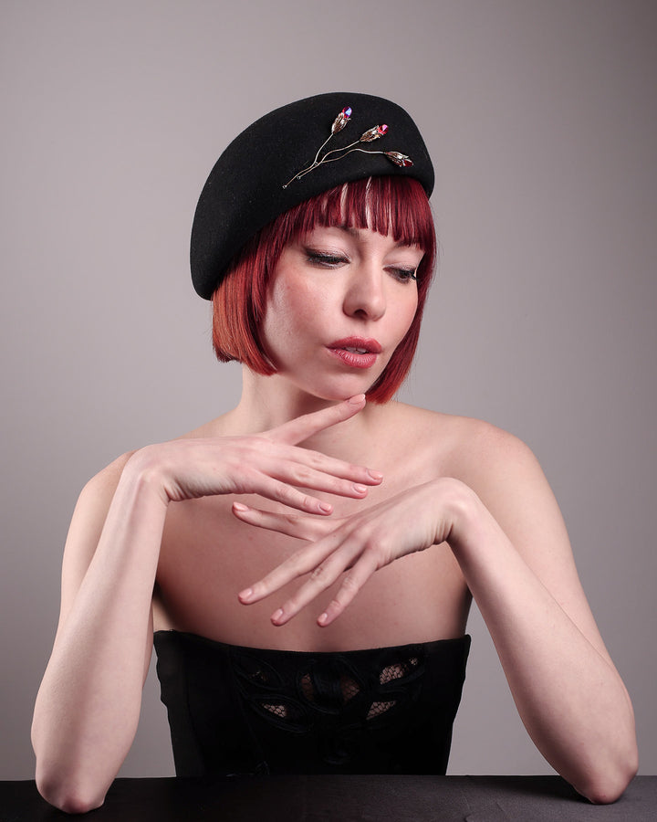 The female model is wearing a wool beret-style hat, showcasing the right front view it focusing to the angle. The black hat is decorated with a real gold-plated brass metal branch motif adorned with crystal-tipped buds. The side beret is higher on the left side and shorter on the right side, and is suggested to be worn tilted. Designed by Merve Bayindir.