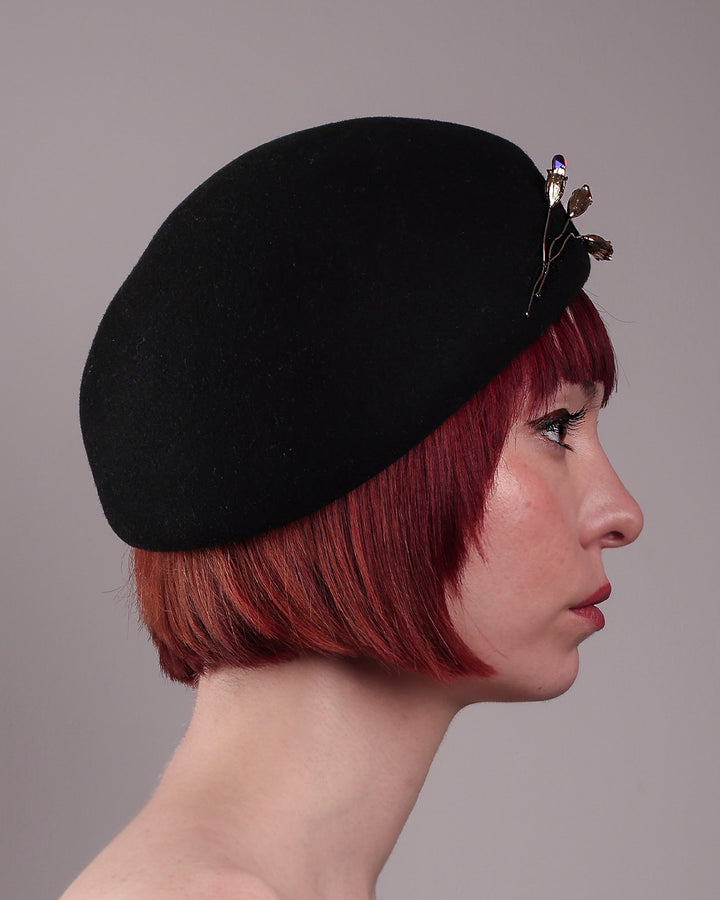 The female model is showcasing the right side close up view of the wool beret-style hat. The black hat is decorated with a real gold-plated brass metal branch motif adorned with crystal-tipped buds. The side beret is higher on the left side and shorter on the right side, and is suggested to be worn tilted. Designed by Merve Bayindir.