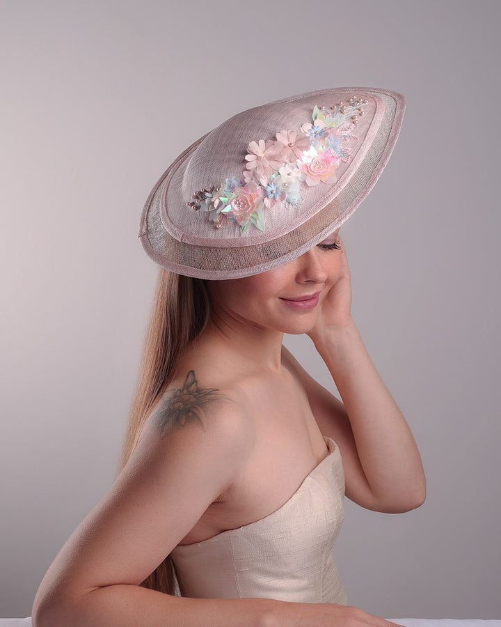 The model is showcasing the front right side from above of the asymetric tear-drop shaped layered pink saucer hat which has two layers of grey in the bottom and topped with a two layer pink colour of same style saucer in a smaller size. The top of the pink layer, hat is decorated with a 3D lace embroiderey embellished with silk flowers, pearls beads and sequins.