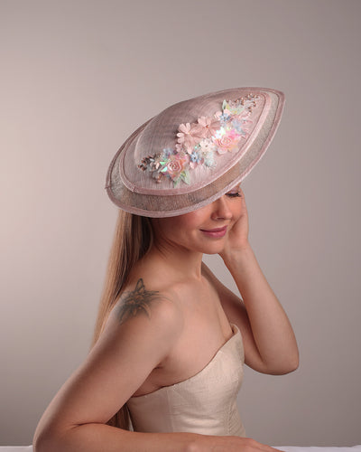 The model is showcasing the front right side from above of the asymetric tear-drop shaped layered pink saucer hat which has two layers of grey in the bottom and topped with a two layer pink colour of same style saucer in a smaller size. The top of the pink layer, hat is decorated with a 3D lace embroiderey embellished with silk flowers, pearls beads and sequins.