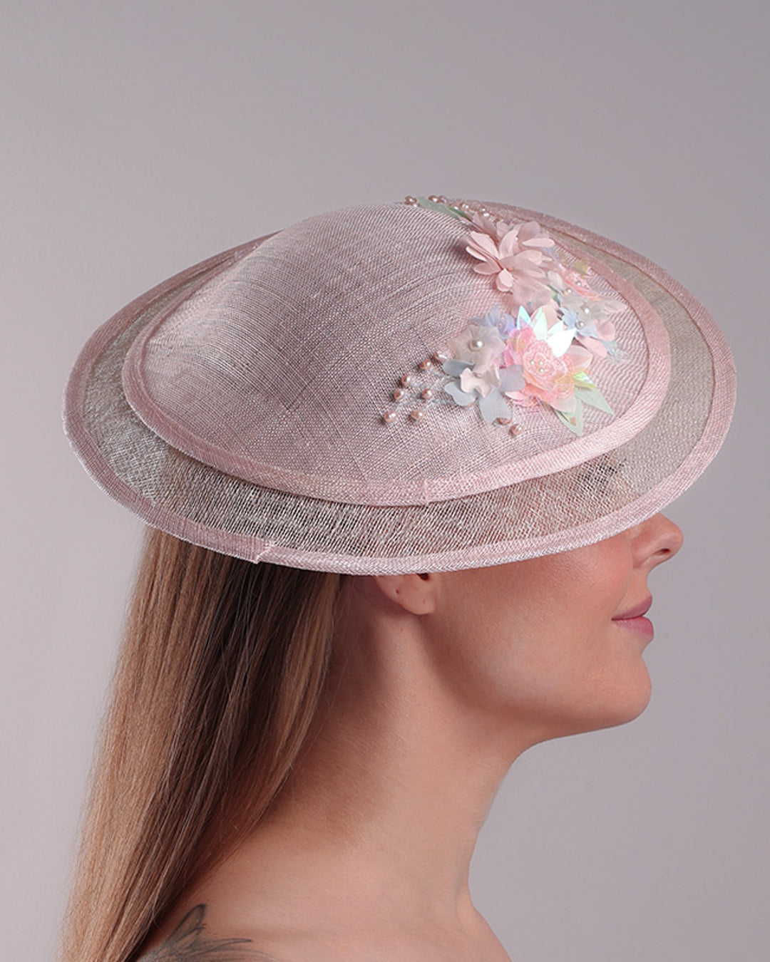 The model is showcasing the right side of the asymetric tear-drop shaped layered pink saucer hat which has two layers of grey in the bottom and topped with a two layer pink colour of same style saucer in a smaller size. The top of the pink layer, hat is decorated with a 3D lace embroiderey embellished with silk flowers, pearls beads and sequins.