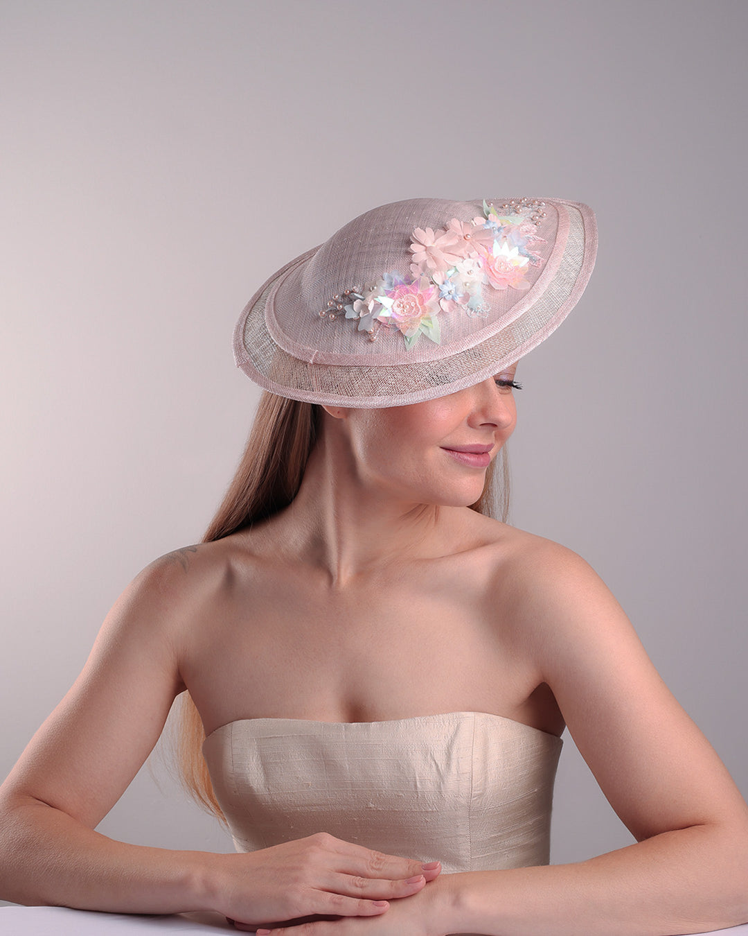 The model is showcasing the front right side from of the asymetric tear-drop shaped layered pink saucer hat which has two layers of grey in the bottom and topped with a two layer pink colour of same style saucer in a smaller size. The top of the pink layer, hat is decorated with a 3D lace embroiderey embellished with silk flowers, pearls beads and sequins.