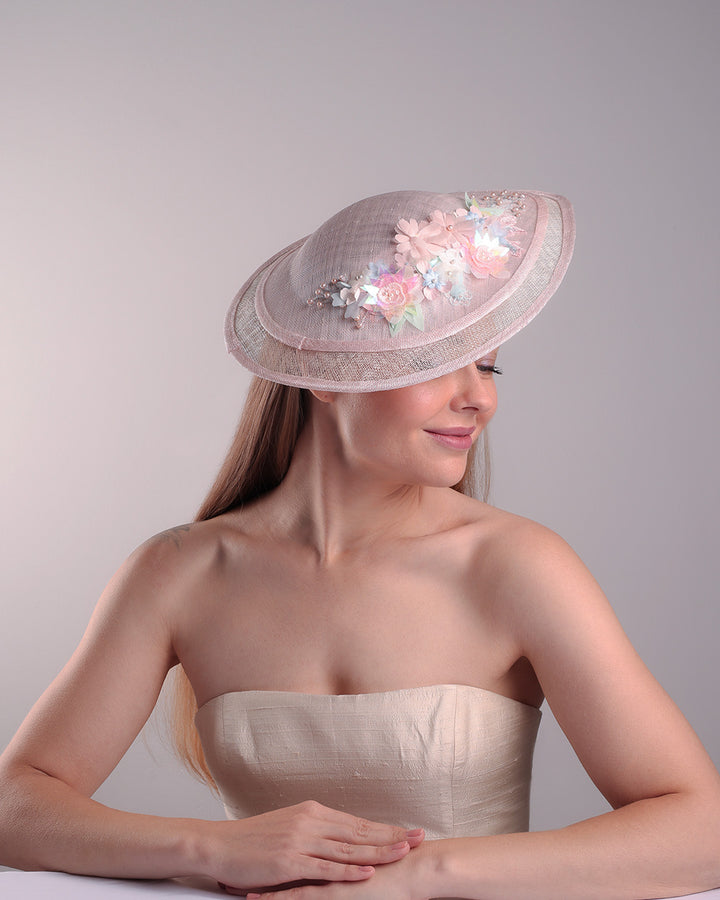 The model is showcasing the front right side from of the asymetric tear-drop shaped layered pink saucer hat which has two layers of grey in the bottom and topped with a two layer pink colour of same style saucer in a smaller size. The top of the pink layer, hat is decorated with a 3D lace embroiderey embellished with silk flowers, pearls beads and sequins.