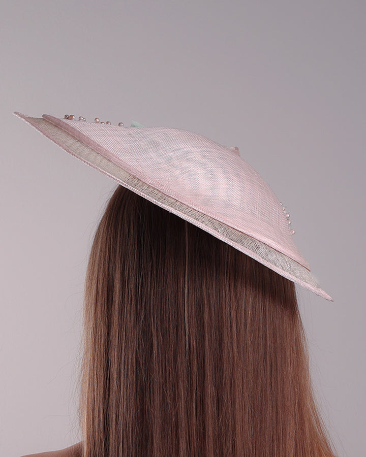 The model is showcasing the back of the asymetric tear-drop shaped layered pink saucer hat which has two layers of grey in the bottom and topped with a two layer pink colour of same style saucer in a smaller size. The top of the pink layer, hat is decorated with a 3D lace embroiderey embellished with silk flowers, pearls beads and sequins.