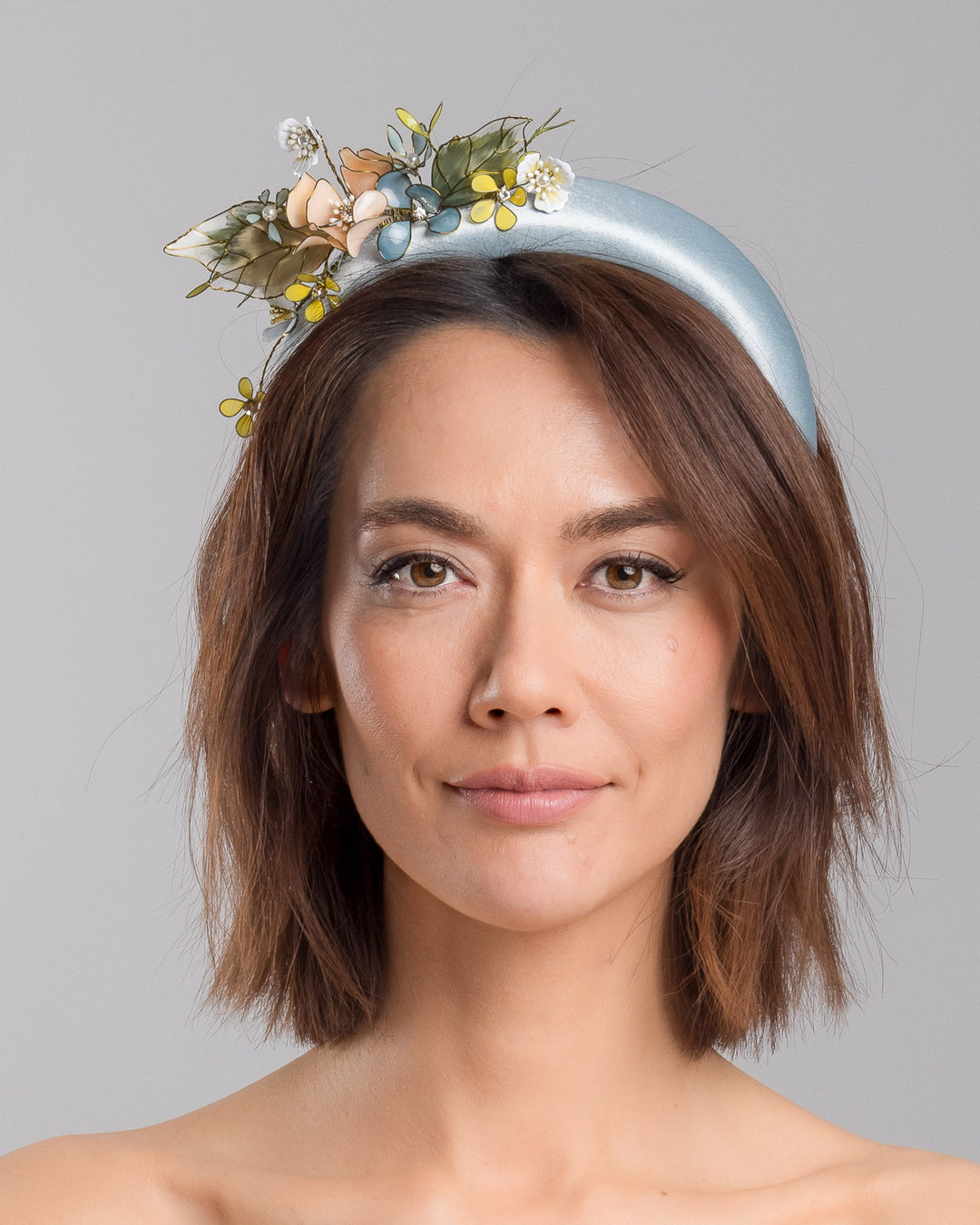 The model is showcasing the front side close up of the powder blue satin padded headband featuring an asymmetrical arrangement of handcrafted translucent flowers and leaves in soft peach, dusty blue, pale yellow, and muted green tones. The petals are delicately edged with fine gold detailing and accented with subtle crystal centers and beadwork, creating an elegant, sculptural statement piece ideal for weddings and refined special occasions. Designed and made by Merve Bayindir Millinery.
