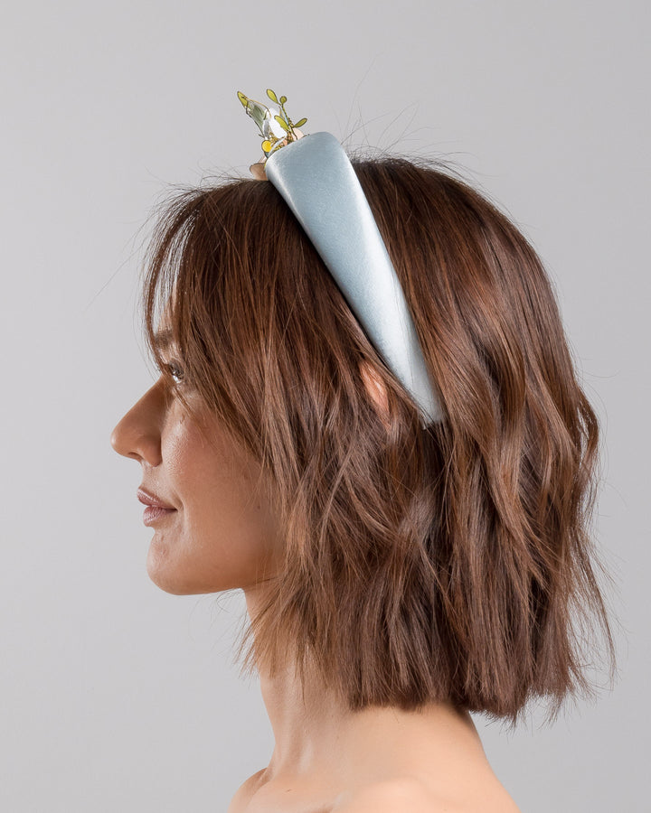 The model is showcasing the left side close up of the powder blue satin padded headband featuring an asymmetrical arrangement of handcrafted translucent flowers and leaves in soft peach, dusty blue, pale yellow, and muted green tones. The petals are delicately edged with fine gold detailing and accented with subtle crystal centers and beadwork, creating an elegant, sculptural statement piece ideal for weddings and refined special occasions. Designed and made by Merve Bayindir Millinery.
