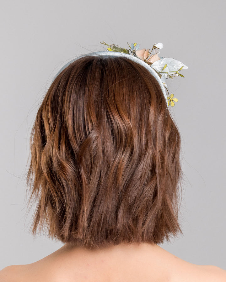 The model is showcasing the back side close up of the powder blue satin padded headband featuring an asymmetrical arrangement of handcrafted translucent flowers and leaves in soft peach, dusty blue, pale yellow, and muted green tones. The petals are delicately edged with fine gold detailing and accented with subtle crystal centers and beadwork, creating an elegant, sculptural statement piece ideal for weddings and refined special occasions. Designed and made by Merve Bayindir Millinery.
