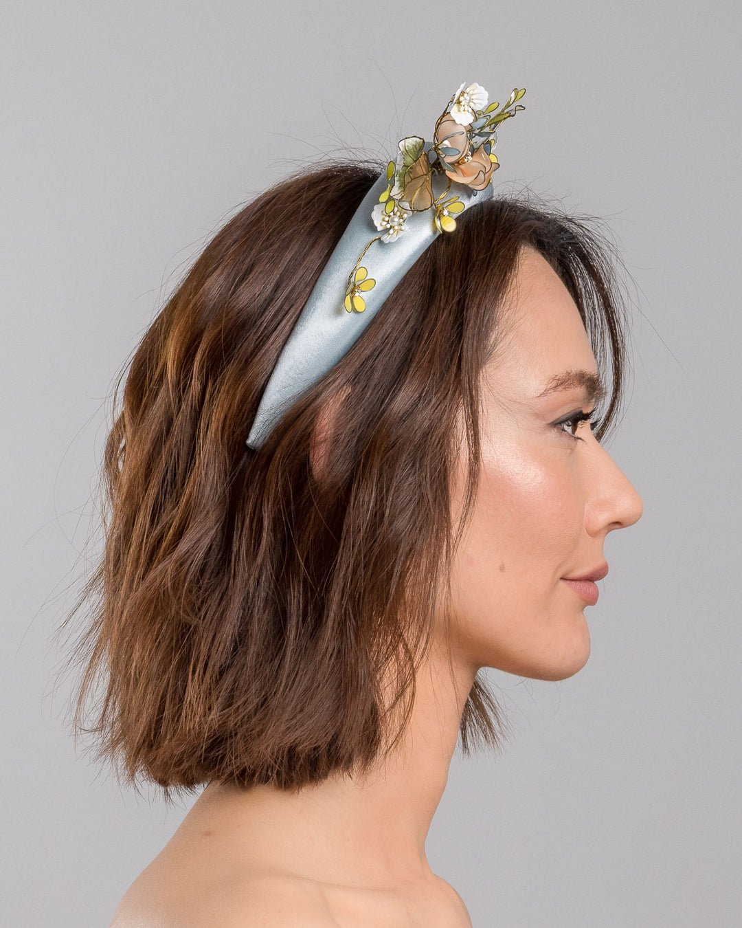 The model is showcasing the right side close up of the powder blue satin padded headband featuring an asymmetrical arrangement of handcrafted translucent flowers and leaves in soft peach, dusty blue, pale yellow, and muted green tones. The petals are delicately edged with fine gold detailing and accented with subtle crystal centers and beadwork, creating an elegant, sculptural statement piece ideal for weddings and refined special occasions. Designed and made by Merve Bayindir Millinery.
