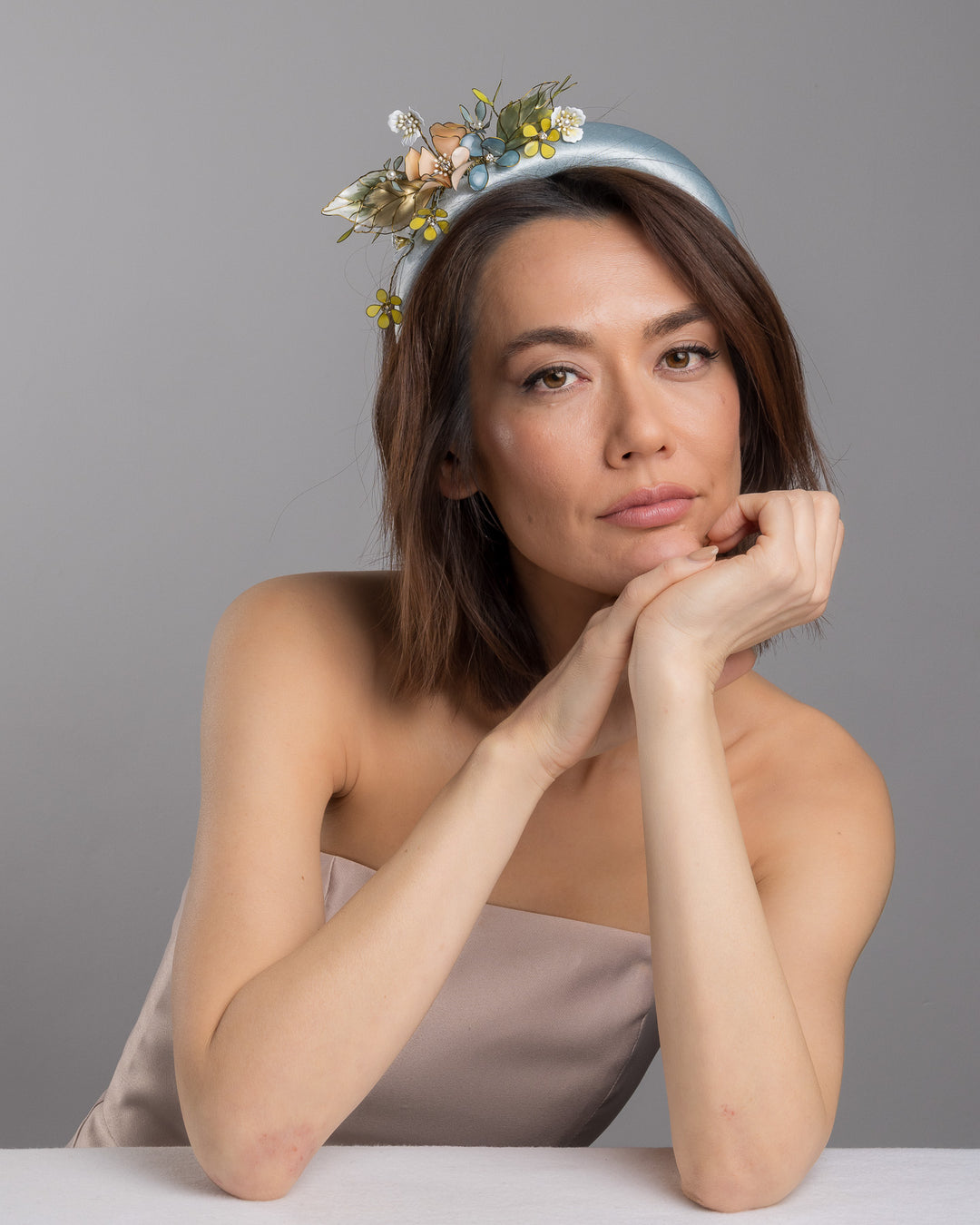 The female model is posing, wearing Selan, the powder blue satin padded headband featuring an asymmetrical arrangement of handcrafted translucent flowers and leaves in soft peach, dusty blue, pale yellow, and muted green tones. The petals are delicately edged with fine gold detailing and accented with subtle crystal centers and beadwork, creating an elegant, sculptural statement piece ideal for weddings and refined special occasions. Designed and made by Merve Bayindir Millinery.
