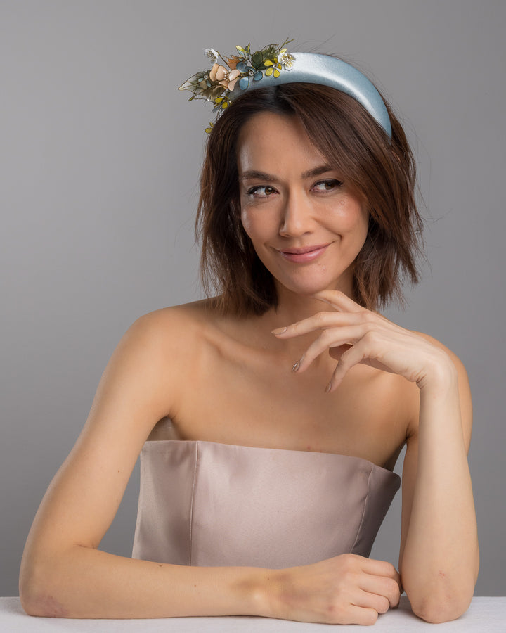 The female model is posing, wearing Selan, the powder blue satin padded headband featuring an asymmetrical arrangement of handcrafted translucent flowers and leaves in soft peach, dusty blue, pale yellow, and muted green tones. The petals are delicately edged with fine gold detailing and accented with subtle crystal centers and beadwork, creating an elegant, sculptural statement piece ideal for weddings and refined special occasions. Designed and made by Merve Bayindir Millinery.
