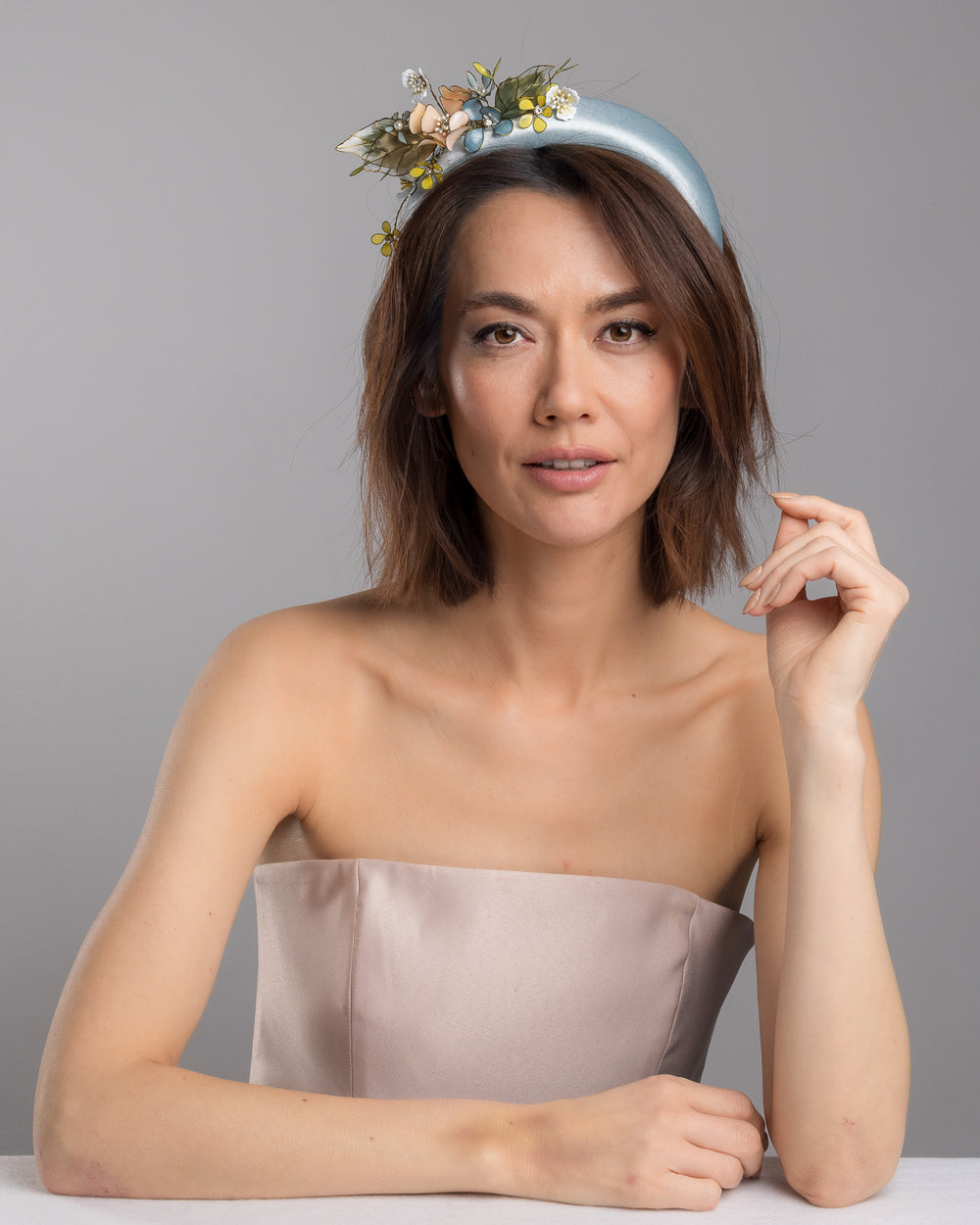 The female model is posing, wearing Selan, the powder blue satin padded headband featuring an asymmetrical arrangement of handcrafted translucent flowers and leaves in soft peach, dusty blue, pale yellow, and muted green tones. The petals are delicately edged with fine gold detailing and accented with subtle crystal centers and beadwork, creating an elegant, sculptural statement piece ideal for weddings and refined special occasions. Designed and made by Merve Bayindir Millinery.

