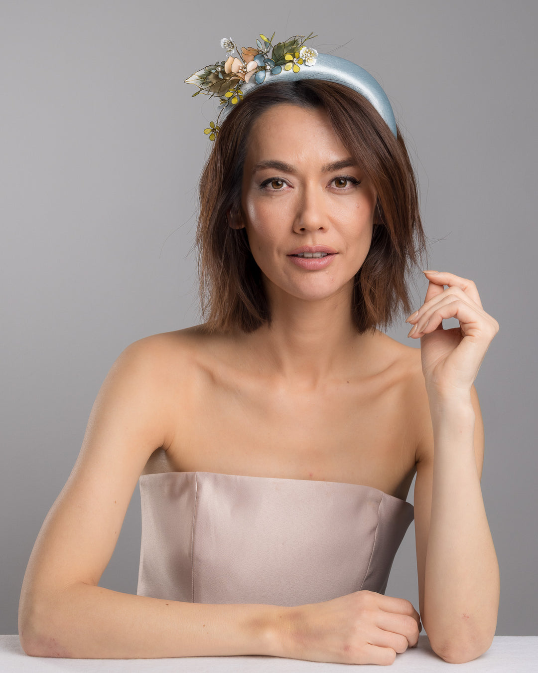 The female model is posing, wearing Selan, the powder blue satin padded headband featuring an asymmetrical arrangement of handcrafted translucent flowers and leaves in soft peach, dusty blue, pale yellow, and muted green tones. The petals are delicately edged with fine gold detailing and accented with subtle crystal centers and beadwork, creating an elegant, sculptural statement piece ideal for weddings and refined special occasions. Designed and made by Merve Bayindir Millinery.
