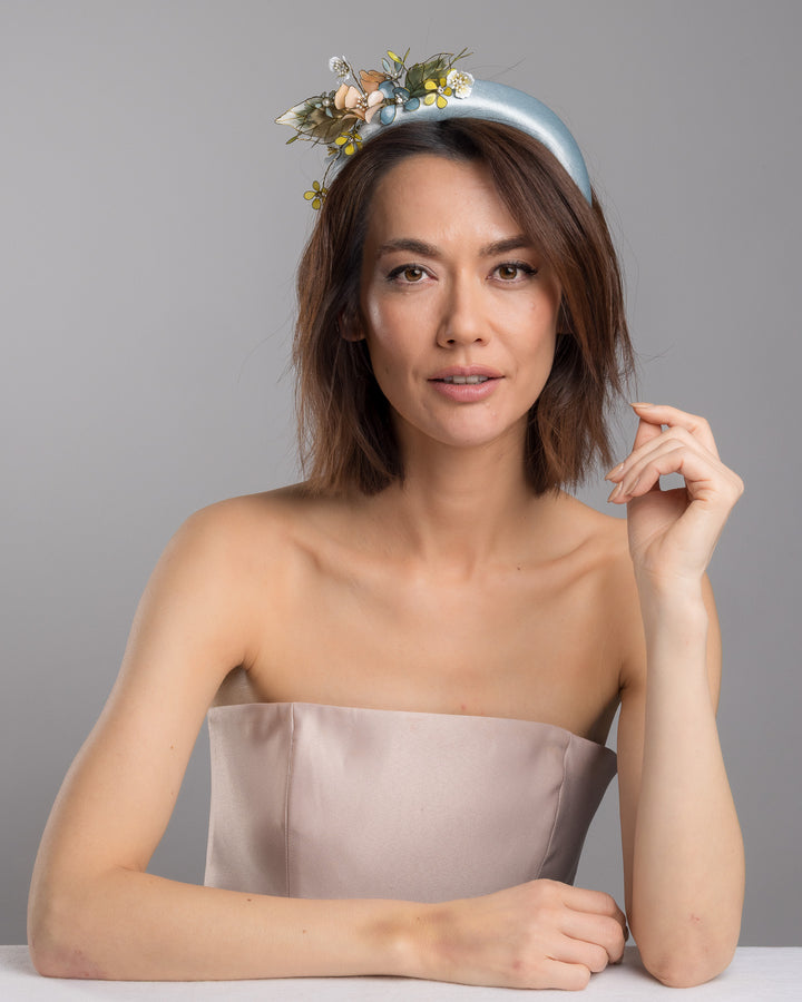 The female model is posing, wearing Selan, the powder blue satin padded headband featuring an asymmetrical arrangement of handcrafted translucent flowers and leaves in soft peach, dusty blue, pale yellow, and muted green tones. The petals are delicately edged with fine gold detailing and accented with subtle crystal centers and beadwork, creating an elegant, sculptural statement piece ideal for weddings and refined special occasions. Designed and made by Merve Bayindir Millinery.
