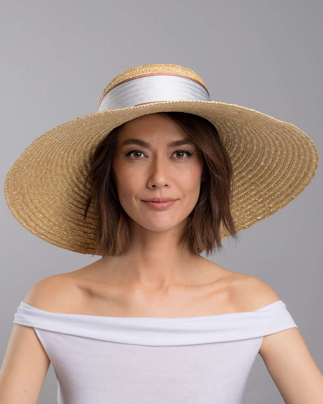 The model is showcasing the front side close up of the natural woven straw hat with a wide brim, wrapped in a taupe and rust silk ribbon and finished with long scarf ties featuring delicate illustrated figures. Designed and made by Merve Bayindir Millinery.
