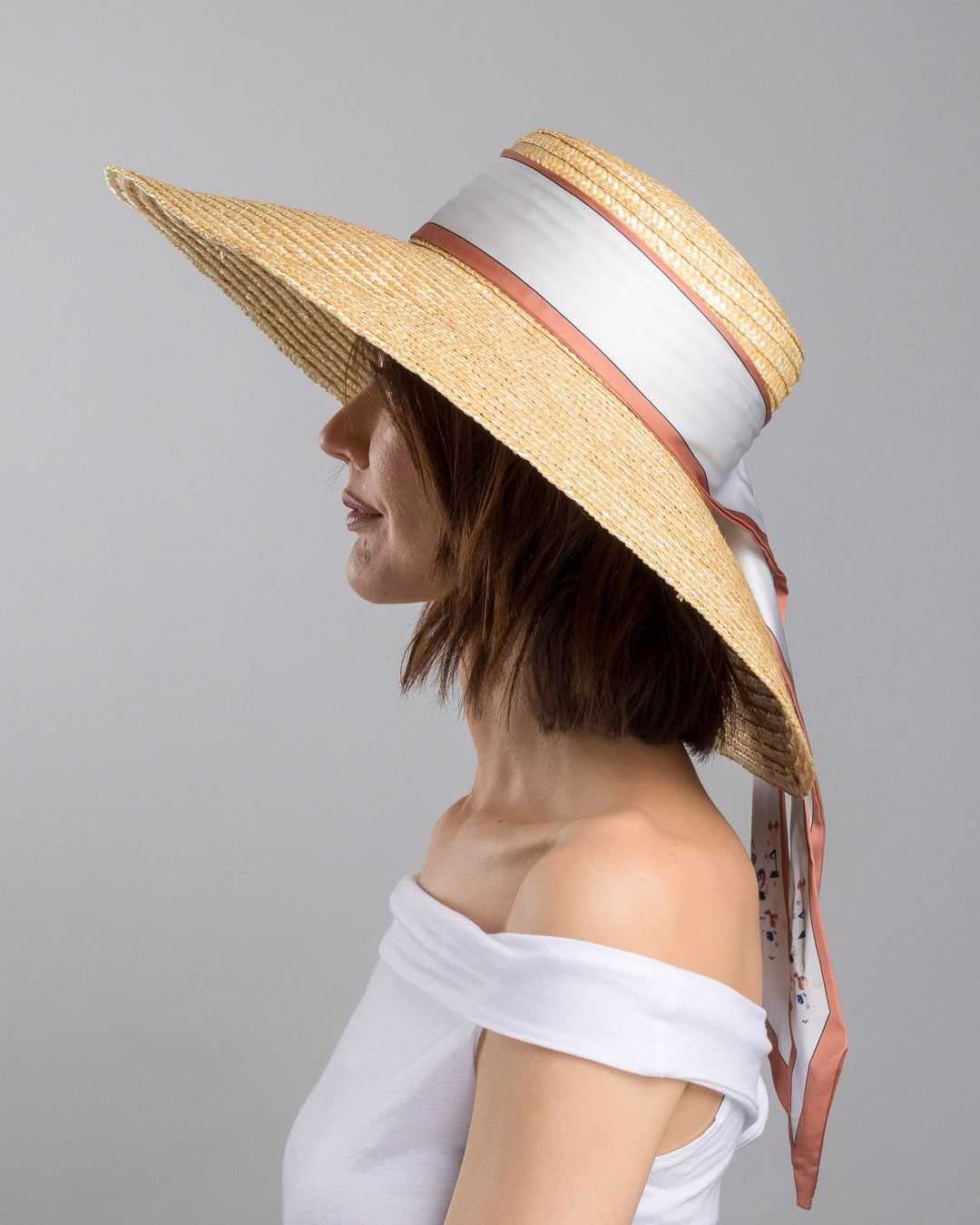 The model is showcasing the left side close up of the natural woven straw hat with a wide brim, wrapped in a taupe and rust silk ribbon and finished with long scarf ties featuring delicate illustrated figures. Designed and made by Merve Bayindir Millinery.
