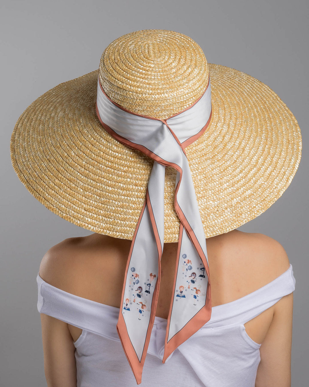 The model is showcasing the back side close up of the natural woven straw hat with a wide brim, wrapped in a taupe and rust silk ribbon and finished with long scarf ties featuring delicate illustrated figures. Designed and made by Merve Bayindir Millinery.
