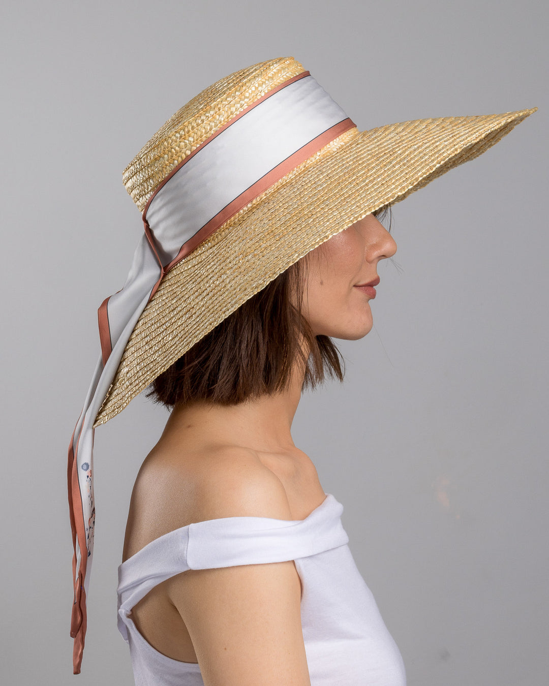 The model is showcasing the right side close up of the natural woven straw hat with a wide brim, wrapped in a taupe and rust silk ribbon and finished with long scarf ties featuring delicate illustrated figures. Designed and made by Merve Bayindir Millinery.

