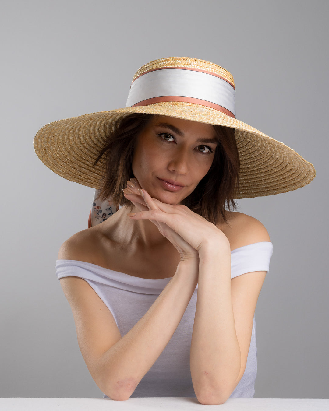 The female model is posing, wearing Sereen, the natural woven straw hat with a wide brim, wrapped in a taupe and rust silk ribbon and finished with long scarf ties featuring delicate illustrated figures. Designed and made by Merve Bayindir Millinery.
