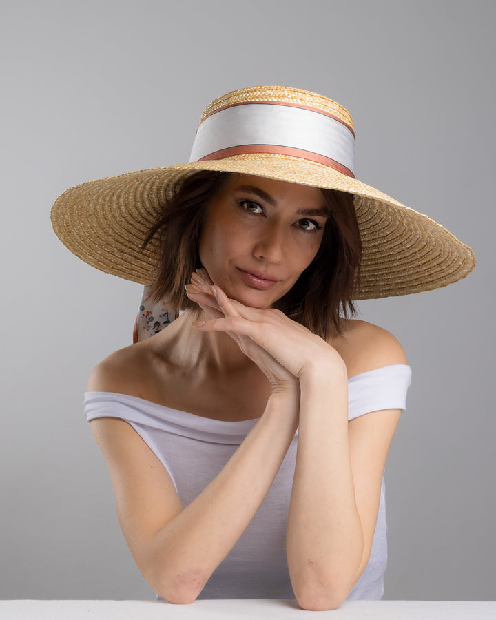 The female model is posing, wearing Sereen, the natural woven straw hat with a wide brim, wrapped in a taupe and rust silk ribbon and finished with long scarf ties featuring delicate illustrated figures. Designed and made by Merve Bayindir Millinery.
