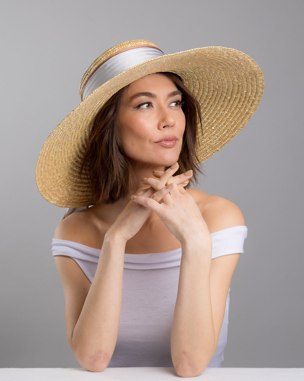 The female model is posing, wearing Sereen, the natural woven straw hat with a wide brim, wrapped in a taupe and rust silk ribbon and finished with long scarf ties featuring delicate illustrated figures. Designed and made by Merve Bayindir Millinery.
