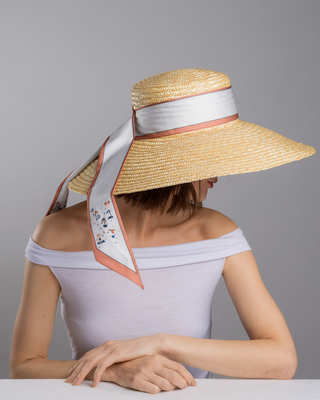 The female model is posing, wearing Sereen, the natural woven straw hat with a wide brim, wrapped in a taupe and rust silk ribbon and finished with long scarf ties featuring delicate illustrated figures. Designed and made by Merve Bayindir Millinery.
