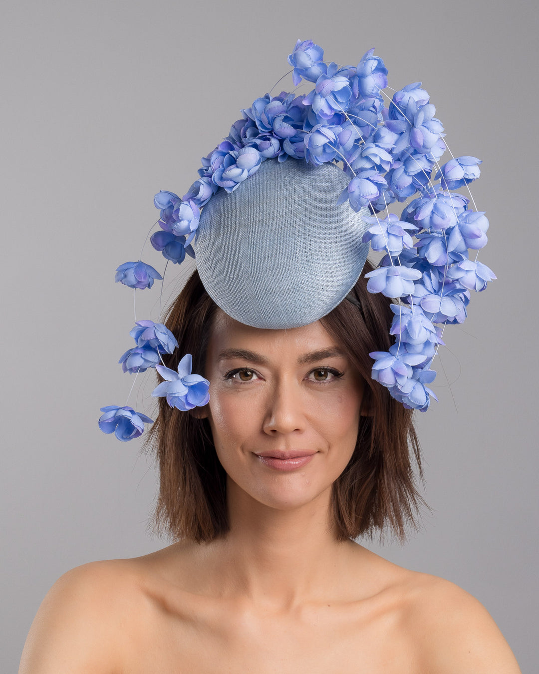 The model is showcasing the front side close up of the light blue sinamay base fascinator with a softly domed shape and high back, adorned with clusters of delicate periwinkle-blue flowers that cascade outward on fine curved stems for an airy, sculptural effect. Designed and made by Merve Bayindir Millinery.
