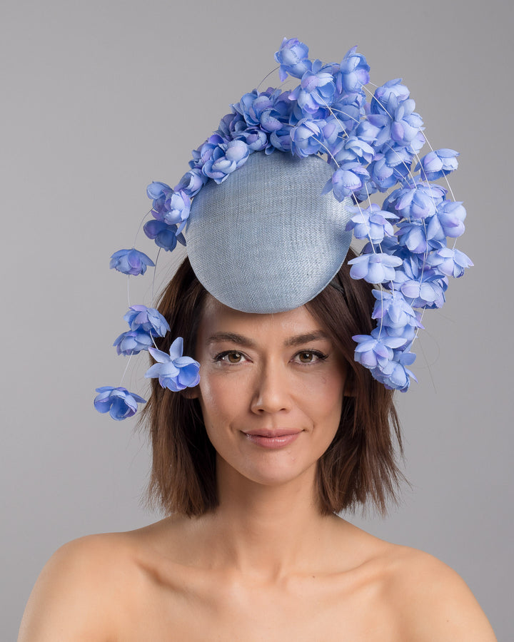 The model is showcasing the front side close up of the light blue sinamay base fascinator with a softly domed shape and high back, adorned with clusters of delicate periwinkle-blue flowers that cascade outward on fine curved stems for an airy, sculptural effect. Designed and made by Merve Bayindir Millinery.
