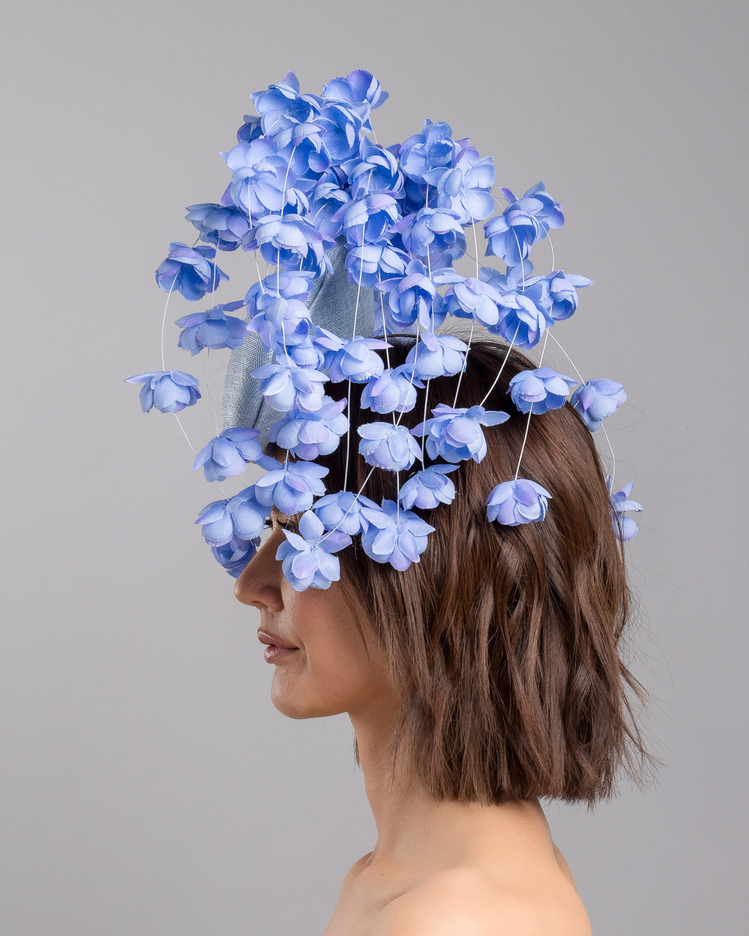 The model is showcasing the left side close up of the light blue sinamay base fascinator with a softly domed shape and high back, adorned with clusters of delicate periwinkle-blue flowers that cascade outward on fine curved stems for an airy, sculptural effect. Designed and made by Merve Bayindir Millinery.

