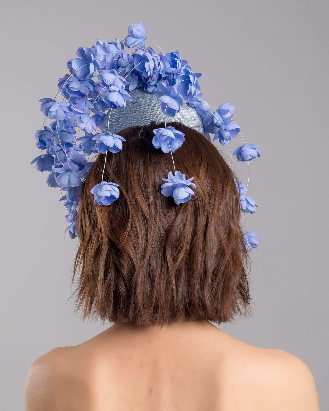 The model is showcasing the back side close up of the light blue sinamay base fascinator with a softly domed shape and high back, adorned with clusters of delicate periwinkle-blue flowers that cascade outward on fine curved stems for an airy, sculptural effect. Designed and made by Merve Bayindir Millinery.

