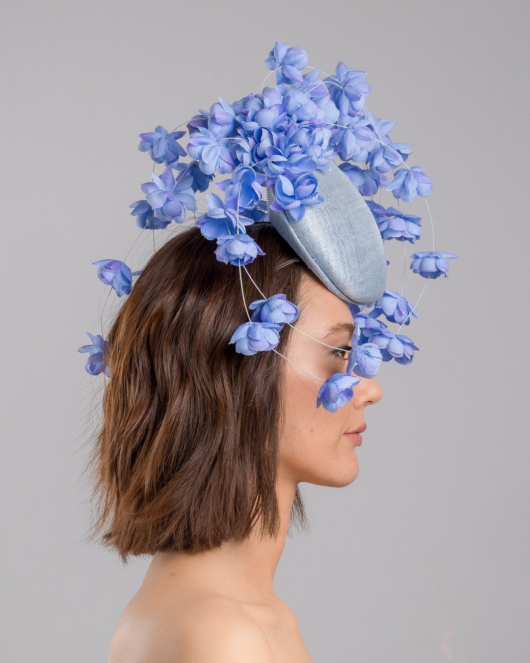 The model is showcasing the right side close up of the light blue sinamay base fascinator with a softly domed shape and high back, adorned with clusters of delicate periwinkle-blue flowers that cascade outward on fine curved stems for an airy, sculptural effect. Designed and made by Merve Bayindir Millinery.
