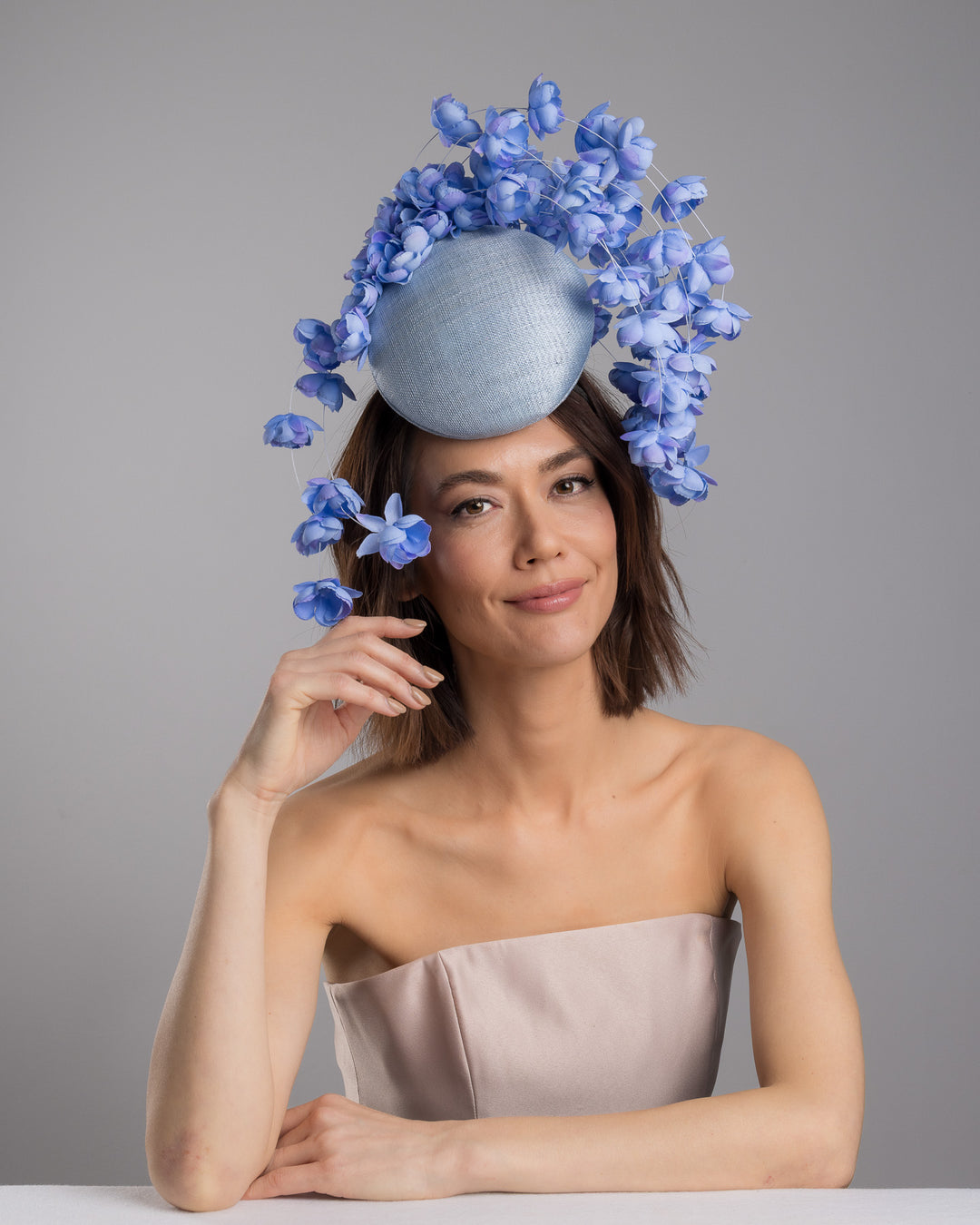 The female model is posing, wearing
Solai, the light blue sinamay base fascinator with a softly domed shape and high back, adorned with clusters of delicate periwinkle-blue flowers that cascade outward on fine curved stems for an airy, sculptural effect. Designed and made by Merve Bayindir Millinery.
