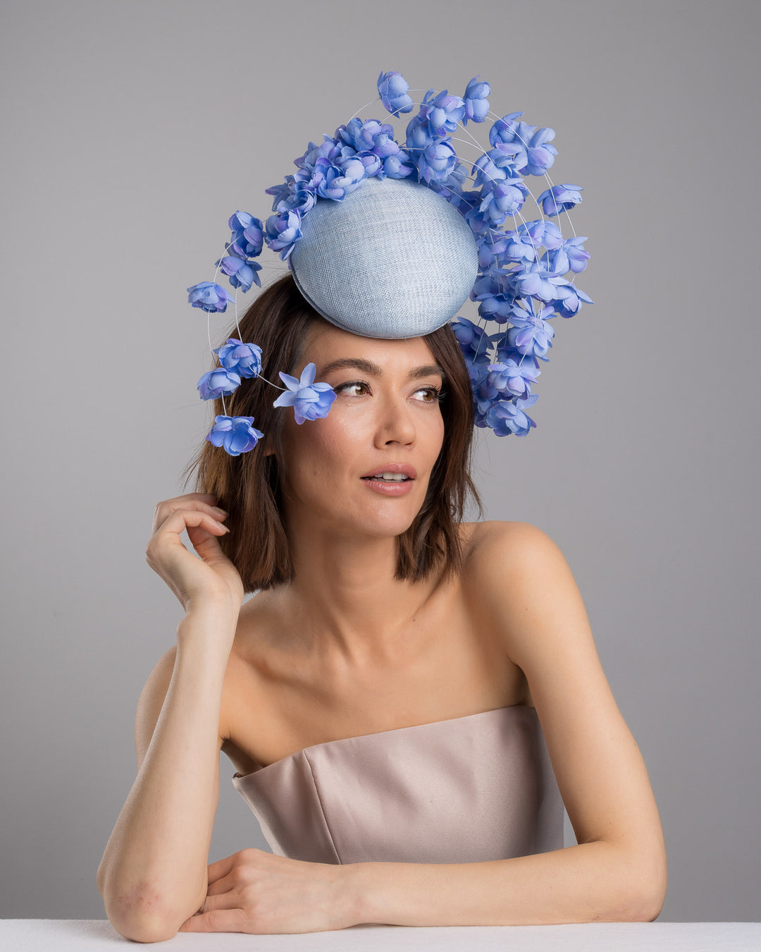 The female model is posing, wearing
Solai, the light blue sinamay base fascinator with a softly domed shape and high back, adorned with clusters of delicate periwinkle-blue flowers that cascade outward on fine curved stems for an airy, sculptural effect. Designed and made by Merve Bayindir Millinery.
