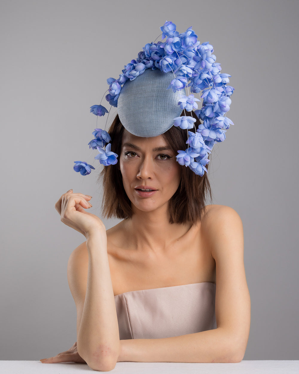 The female model is posing, wearing
Solai, the light blue sinamay base fascinator with a softly domed shape and high back, adorned with clusters of delicate periwinkle-blue flowers that cascade outward on fine curved stems for an airy, sculptural effect. Designed and made by Merve Bayindir Millinery.
