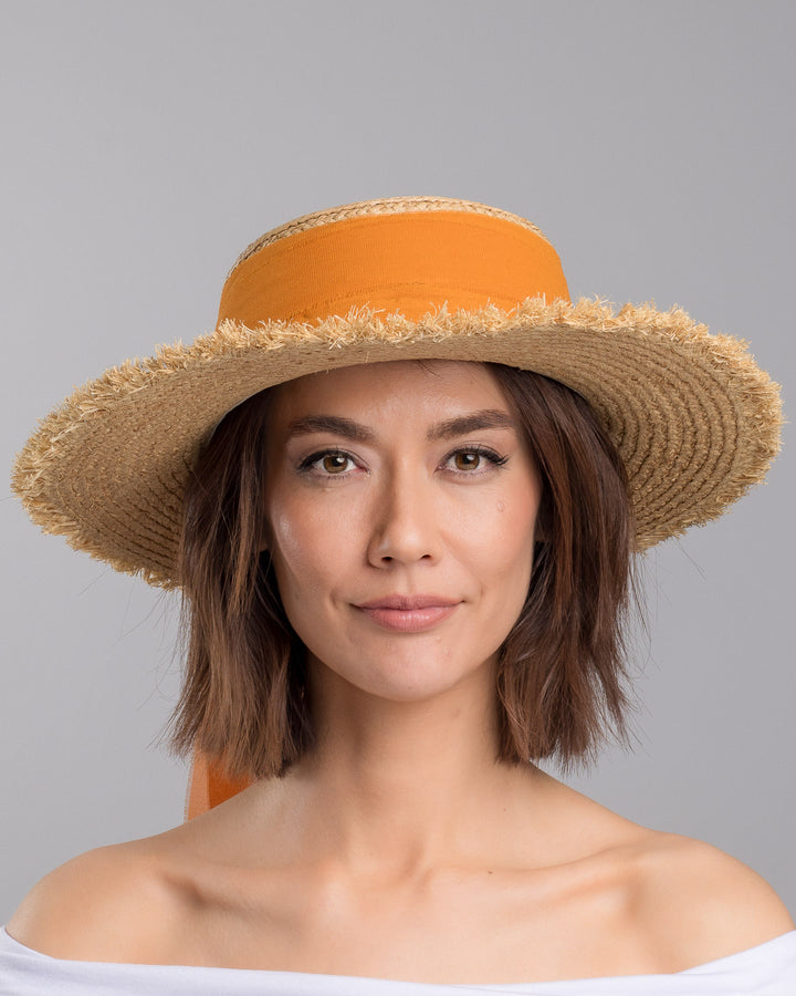 The model is showcasing the front side close up of the natural woven straw boater hat with a wide frayed brim, accented with long sheer orange ribbon ties at the back. Designed and Made by Merve Bayindir millinery.