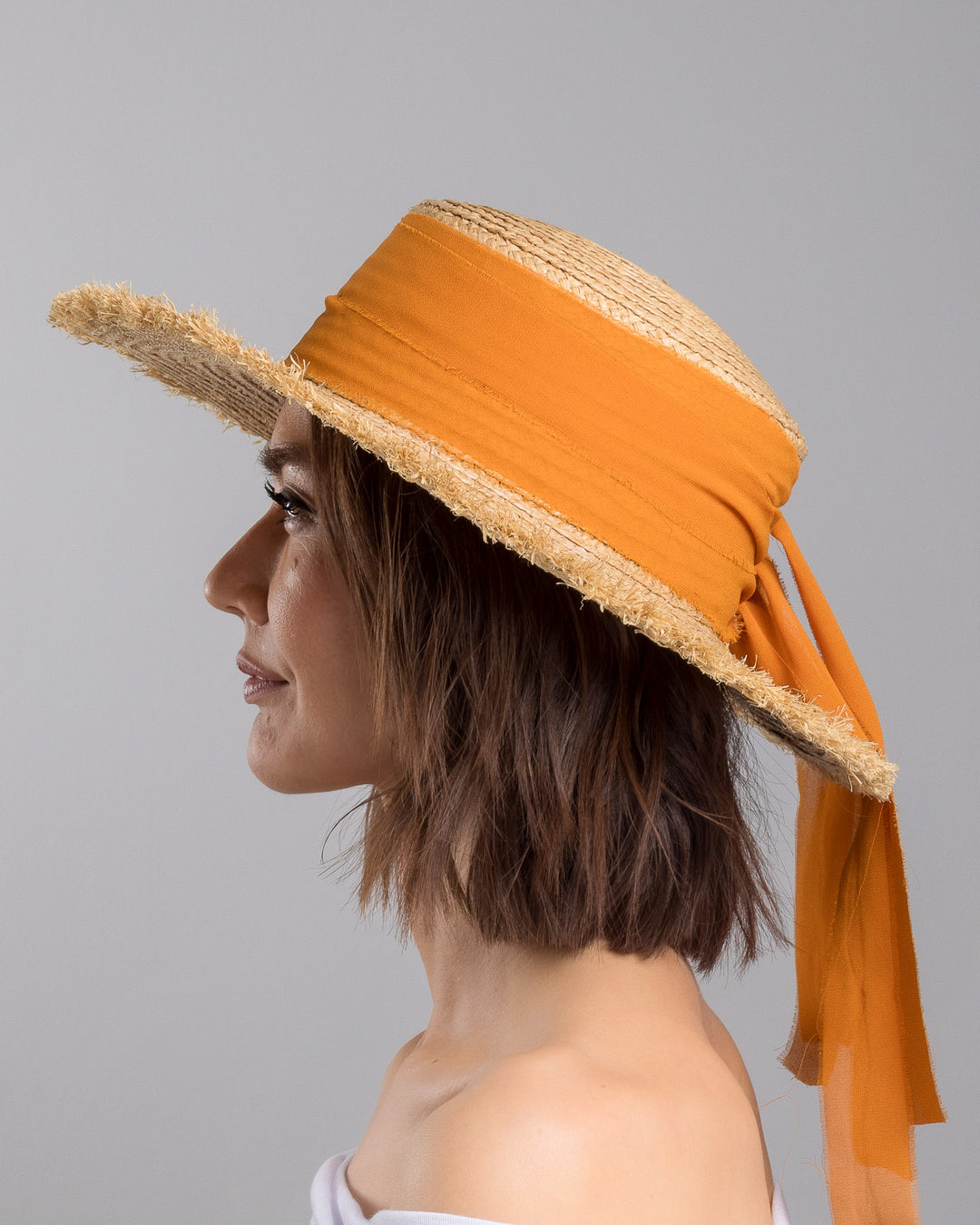 The model is showcasing the left side close up of the natural woven straw boater hat with a wide frayed brim, accented with long sheer orange ribbon ties at the back. Designed and Made by Merve Bayindir millinery.