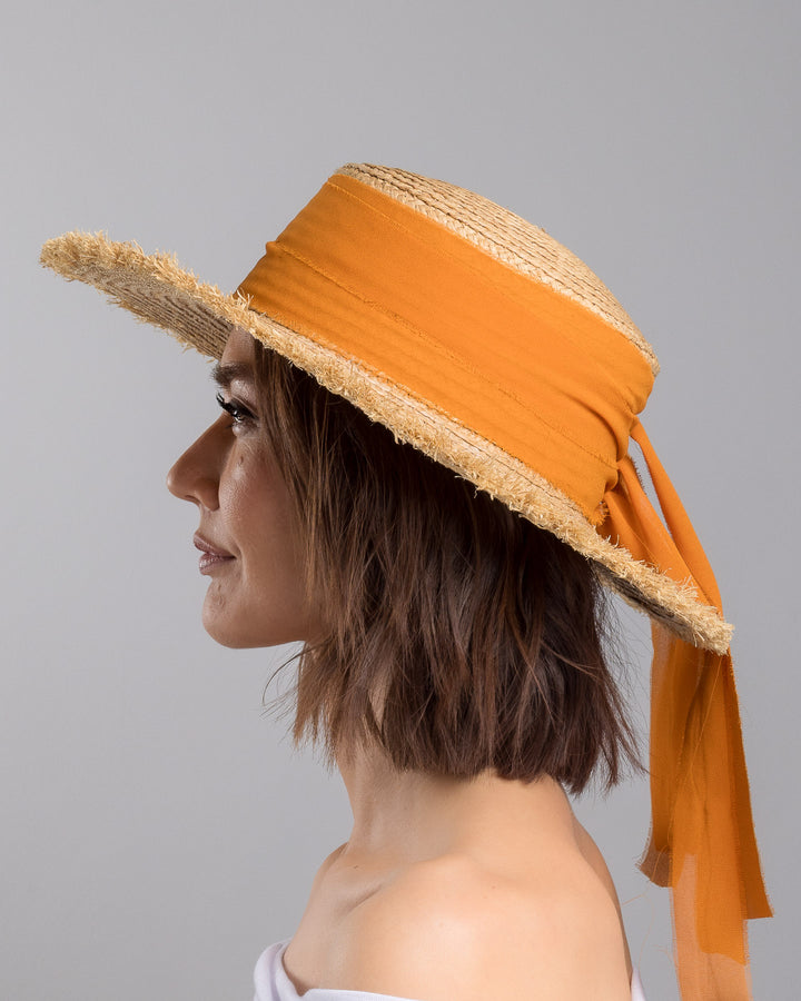 The model is showcasing the left side close up of the natural woven straw boater hat with a wide frayed brim, accented with long sheer orange ribbon ties at the back. Designed and Made by Merve Bayindir millinery.
