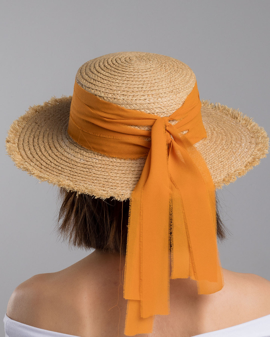 The model is showcasing the back side close up of the natural woven straw boater hat with a wide frayed brim, accented with long sheer orange ribbon ties at the back. Designed and Made by Merve Bayindir millinery.