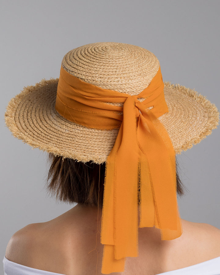 The model is showcasing the back side close up of the natural woven straw boater hat with a wide frayed brim, accented with long sheer orange ribbon ties at the back. Designed and Made by Merve Bayindir millinery.