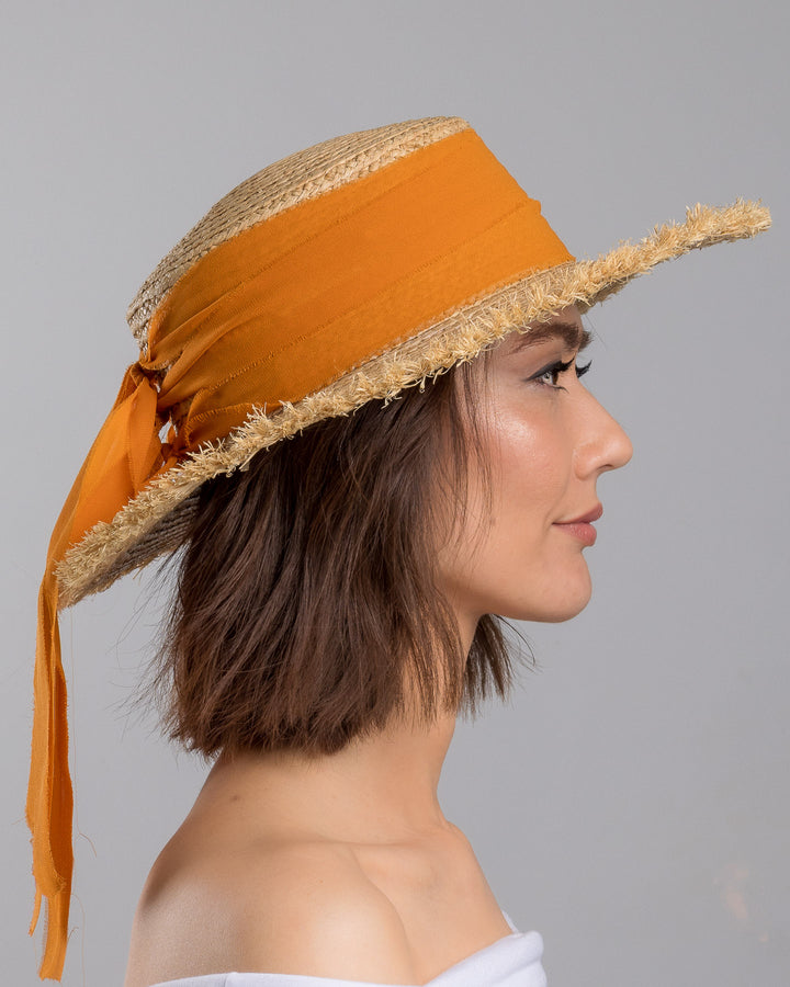 The model is showcasing the right side close up of the natural woven straw boater hat with a wide frayed brim, accented with long sheer orange ribbon ties at the back. Designed and Made by Merve Bayindir millinery.