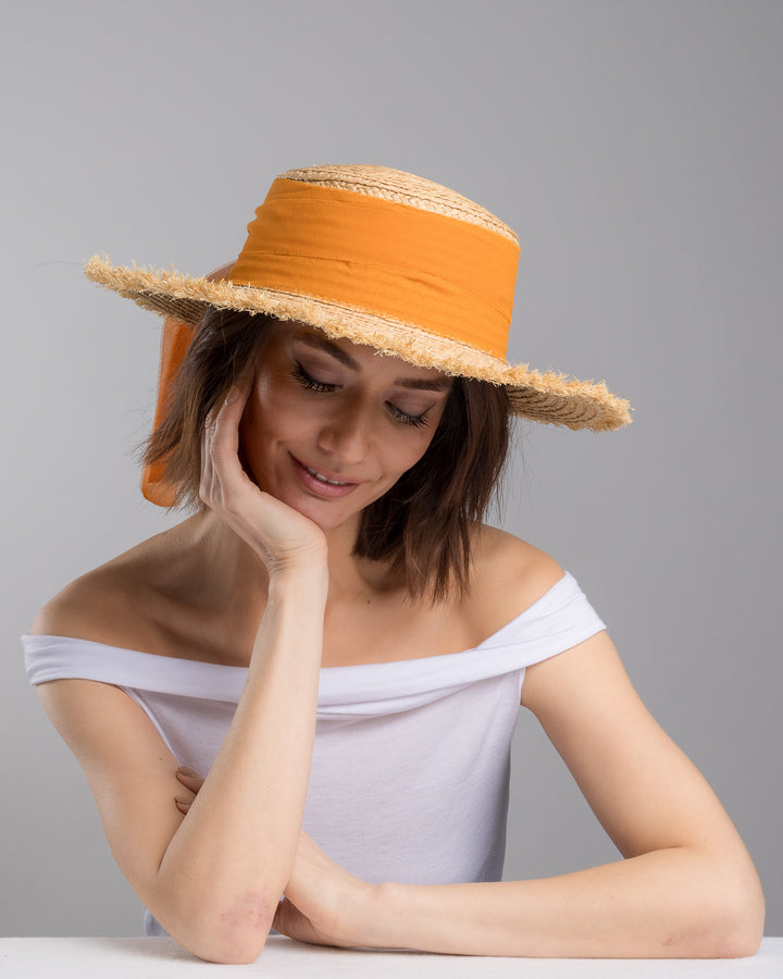 The female model is posing, wearing Solea,  the natural woven straw boater hat with a wide frayed brim, accented with long sheer orange ribbon ties at the back. Designed and Made by Merve Bayindir millinery.