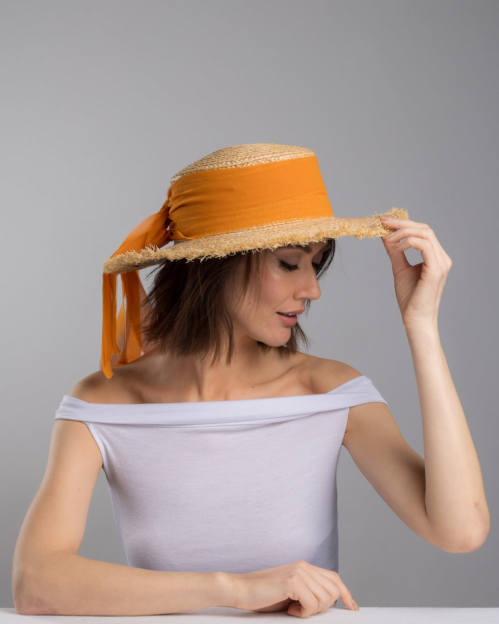 The female model is posing, wearing Solea,  the natural woven straw boater hat with a wide frayed brim, accented with long sheer orange ribbon ties at the back. Designed and Made by Merve Bayindir millinery.