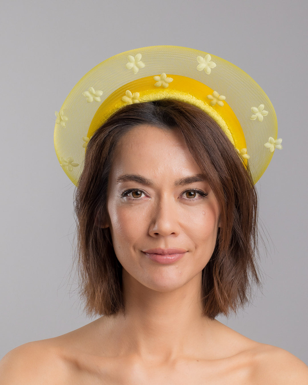 The model is showcasing the front side close up of the lemon yellow headband fascinator featuring a satin-covered base encircled by a sheer structured halo, embellished with small tonal floral appliqués for a light, textured finish. Designed and made by Merve Bayindir Millinery.