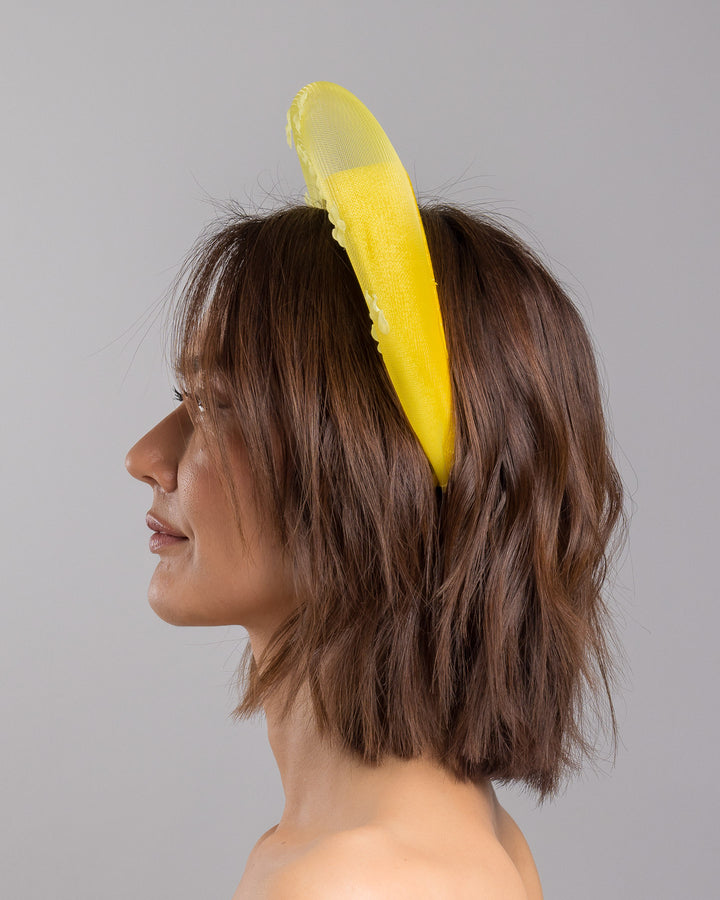 The model is showcasing the left side close up of the lemon yellow headband fascinator featuring a satin-covered base encircled by a sheer structured halo, embellished with small tonal floral appliqués for a light, textured finish. Designed and made by Merve Bayindir Millinery.