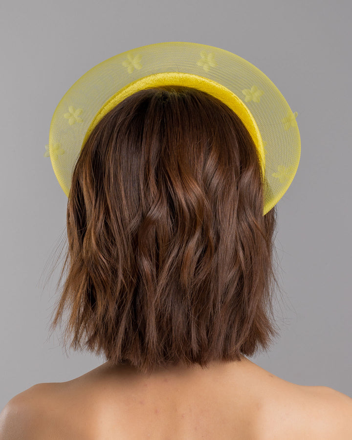 The model is showcasing the back side close up of the lemon yellow headband fascinator featuring a satin-covered base encircled by a sheer structured halo, embellished with small tonal floral appliqués for a light, textured finish. Designed and made by Merve Bayindir Millinery.