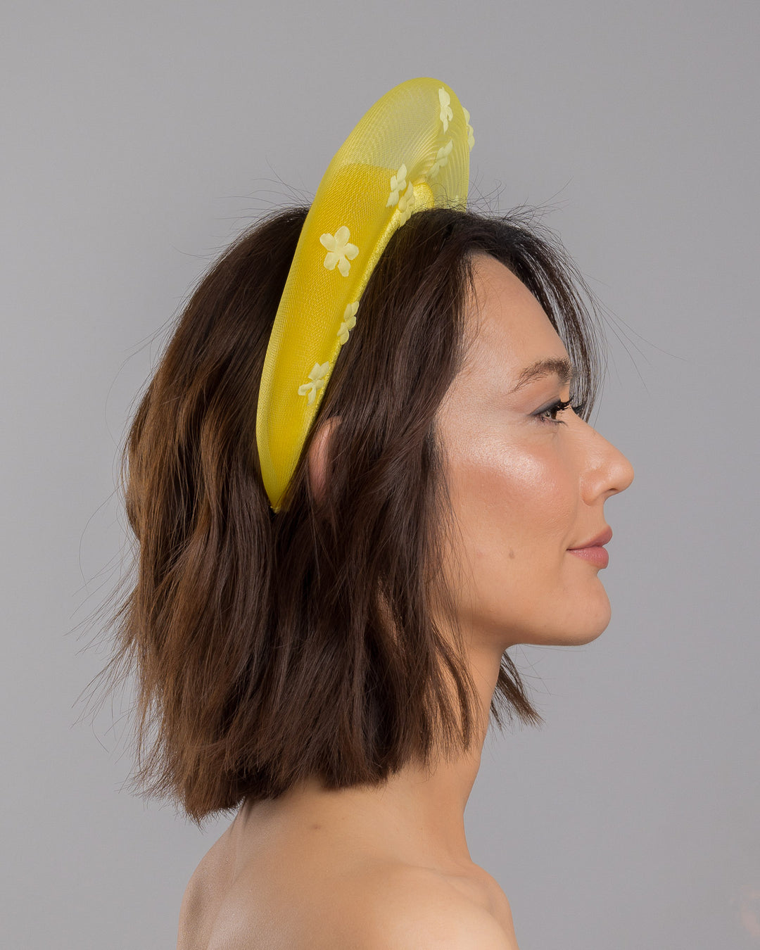 The model is showcasing the right side close up of the lemon yellow headband fascinator featuring a satin-covered base encircled by a sheer structured halo, embellished with small tonal floral appliqués for a light, textured finish. Designed and made by Merve Bayindir Millinery.
