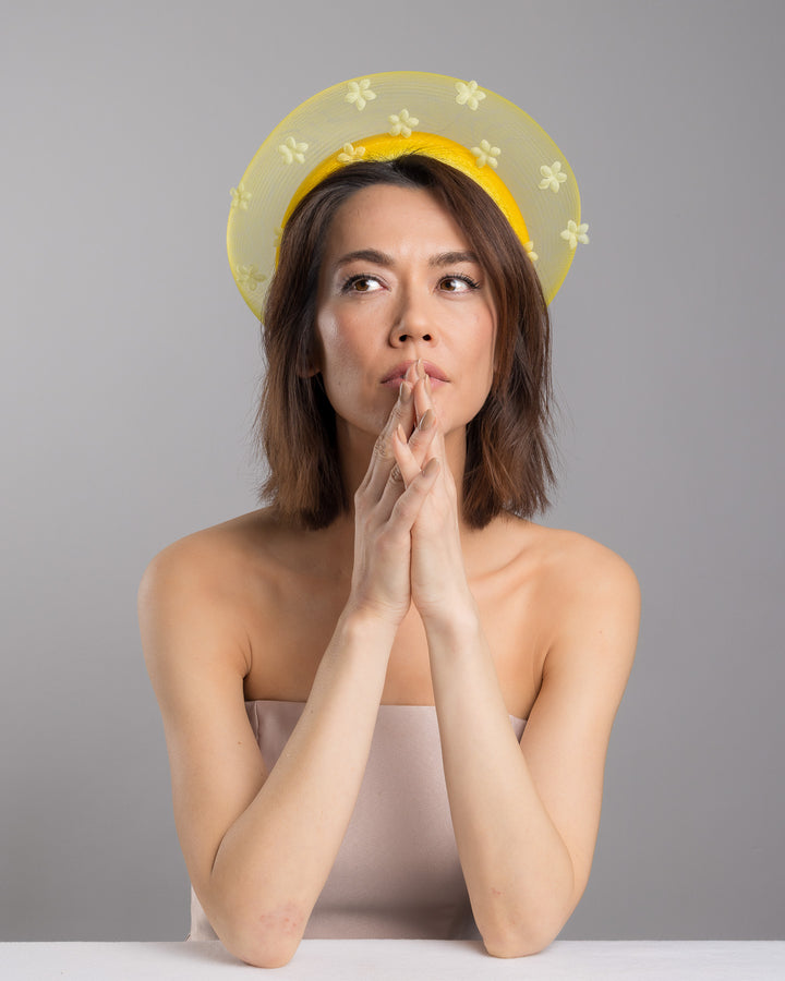 The female model is posing, wearing Soline, the lemon yellow headband fascinator featuring a satin-covered base encircled by a sheer structured halo, embellished with small tonal floral appliqués for a light, textured finish. Designed and made by Merve Bayindir Millinery.